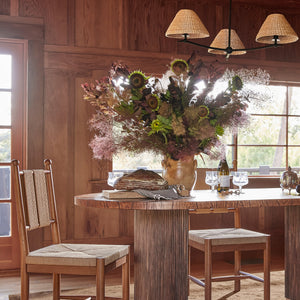 A rustic dining room scene with a large floral arrangement in a vase on a wooden table, with two wooden chairs and stools, a hanging lamp, and wood-paneled walls in the background. - Number 2