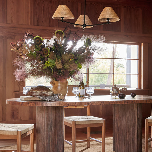 A rustic dining room scene with a round wooden table, four matching wooden chairs with woven seats, a large floral arrangement in a vase, and a chandelier above. The room has wood-paneled walls and a window in the background. - Image 2