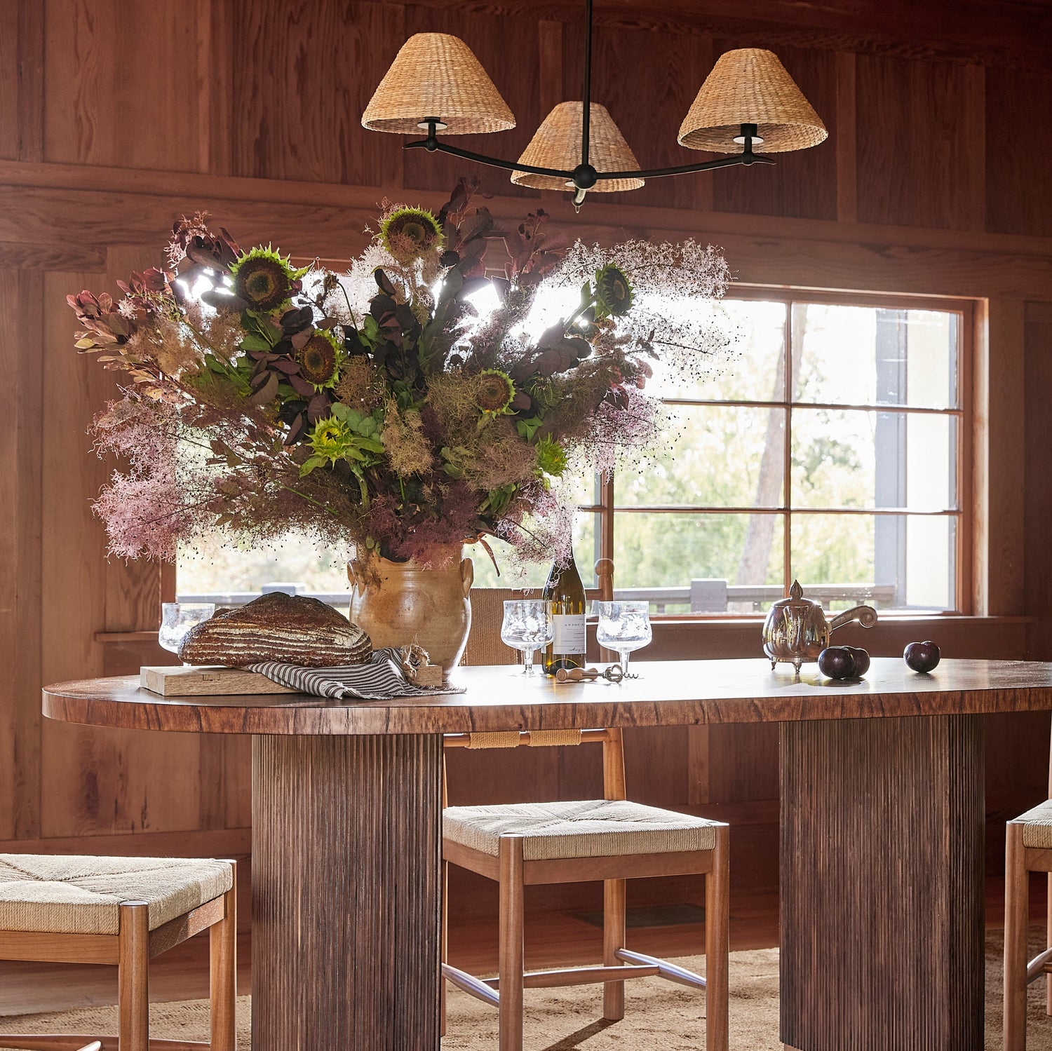 A rustic dining room scene with a round wooden table, four matching wooden chairs with woven seats, a large floral arrangement in a vase, and a chandelier above. The room has wood-paneled walls and a window in the background.- Number 2