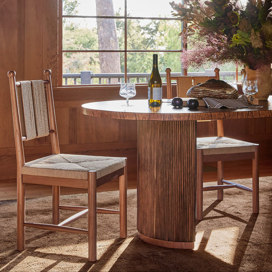 A rustic dining room scene featuring a round wooden table with a fluted pedestal base and two matching wooden chairs with woven seat cushions, set in a warm wood-paneled room with a window and hanging lamps. - Image 2