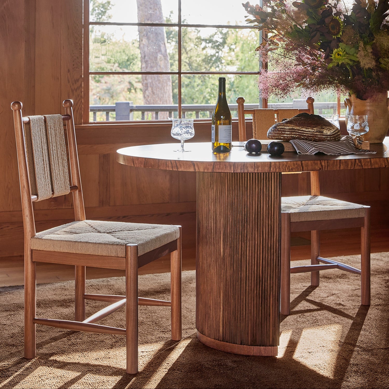 A rustic dining room scene featuring a round wooden table with a fluted pedestal base and two matching wooden chairs with woven seat cushions, set in a warm wood-paneled room with a window and hanging lamps.- Number 10