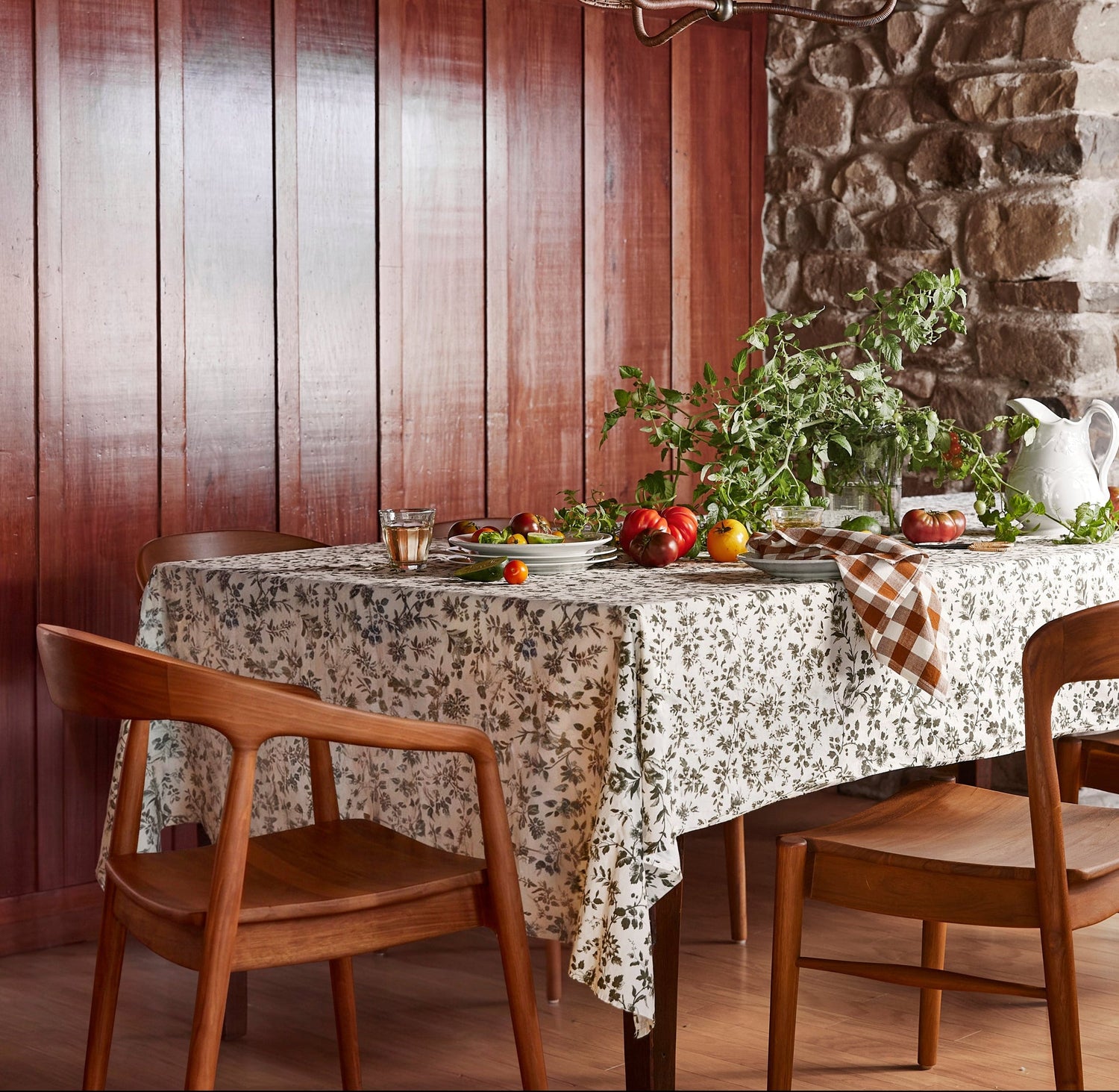 A rustic dining room scene with a floral tablecloth on a long table, surrounded by wooden chairs. The table is set with greenery, tomatoes, and a white pitcher, against a backdrop of dark wood paneling and a stone wall.- Number 2
