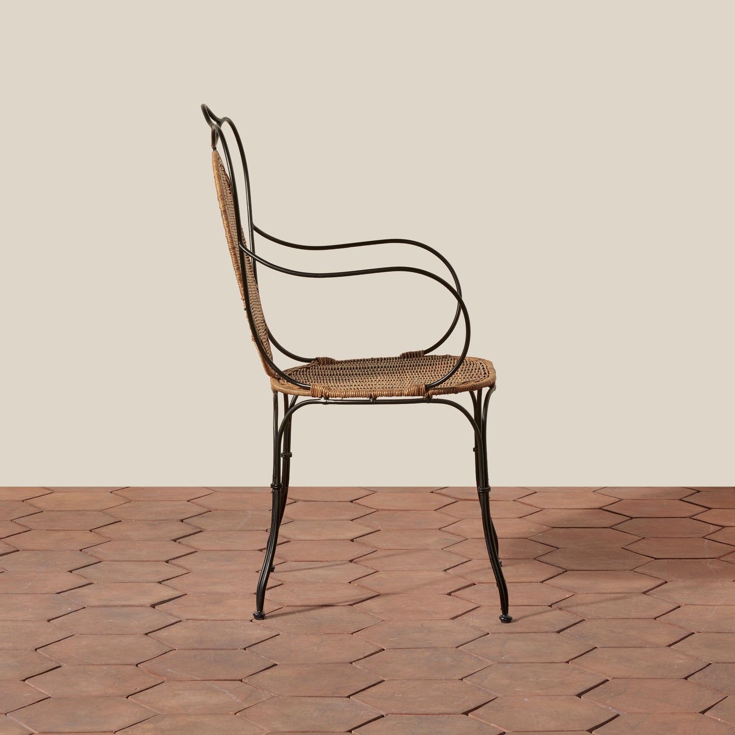 A vintage-style metal and cane armchair shown from the side, positioned on a terracotta tiled floor against a solid beige background.- Number 4