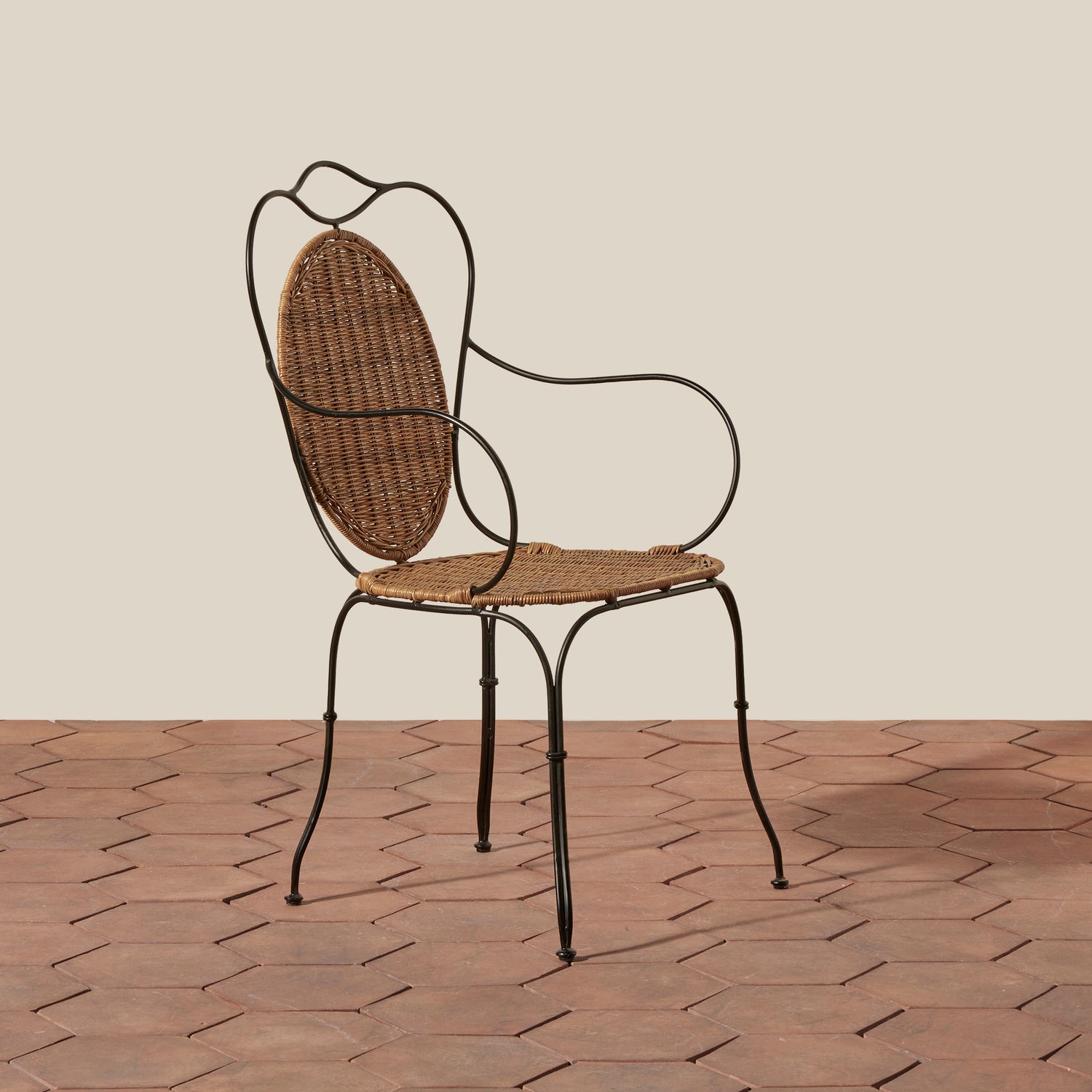 A classic outdoor dining chair with a black metal frame and a woven brown wicker seat and backrest, set against a beige wall and terracotta tiled floor.- Number 1