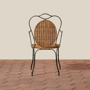 A vintage-style metal and wicker chair with a curved black iron frame, woven oval backrest, and woven seat, set against a beige wall and terracotta tile floor. - Number 6