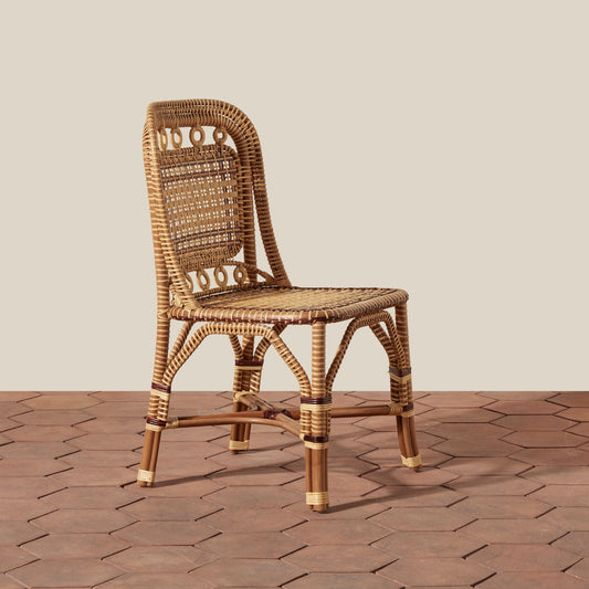 A natural woven rattan dining chair with a rounded backrest and decorative circular woven patterns, set against a neutral beige wall and brown hexagonal tile floor.