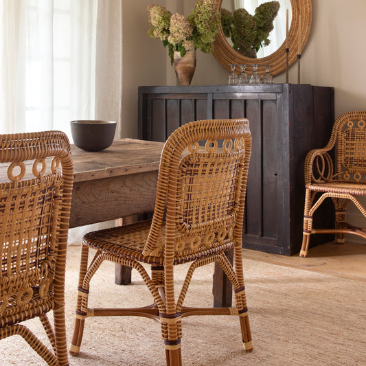 A rustic dining room scene featuring natural woven rattan chairs around a weathered wooden table, with a dark wood cabinet and a round mirror in the background. - Image 2