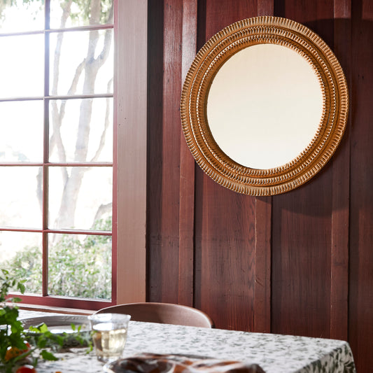 A round wall mirror with an ornate gold-toned mirror hangs on a dark wood-paneled wall above a dining table set with a patterned tablecloth, plates, and a checkered napkin in a sunlit room. - Image 2