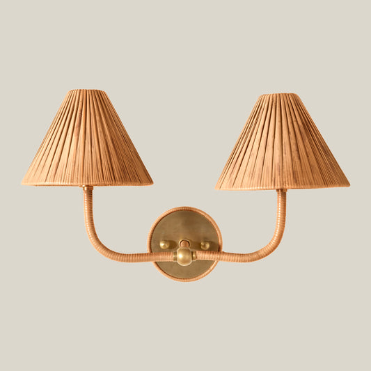 A two-light wall sconce with pleated beige shades and a warm brass metal frame, shown against a solid light gray background.
