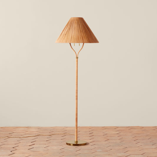 A tall floor lamp with a pleated conical shade, a slender bamboo-like stem, and a round brass base, standing on a terracotta tile floor against a plain beige wall.