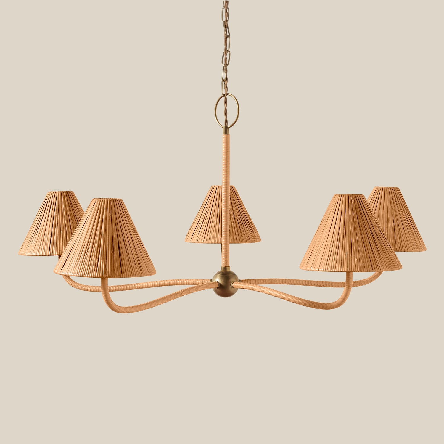 A five-light chandelier with curved brass-toned metal arms and pleated beige fabric shades, suspended from a chain against a solid beige background.- Number 1
