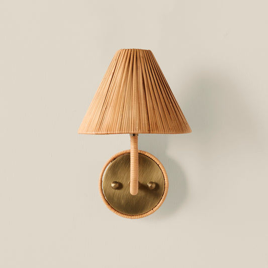 A wall-mounted light fixture with a pleated beige shade, a wrapped beige stem, and a round brass-toned metal backplate, shown against a plain light beige wall.