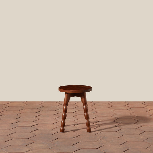 A small round wooden stool with four turned wood legs, shown on a terracotta hexagonal tile floor against a solid beige wall. - Image 2
