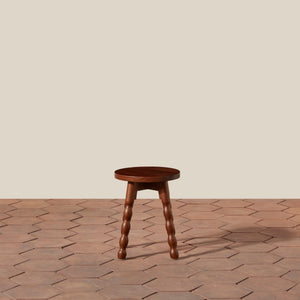 A small round wooden stool with four turned wood legs, shown on a terracotta hexagonal tile floor against a solid beige wall. - Number 2