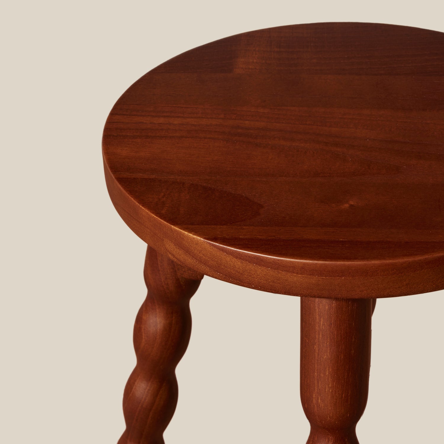 Close-up of a round wooden stool top with turned wood legs, shown against a plain light beige background.- Number 3