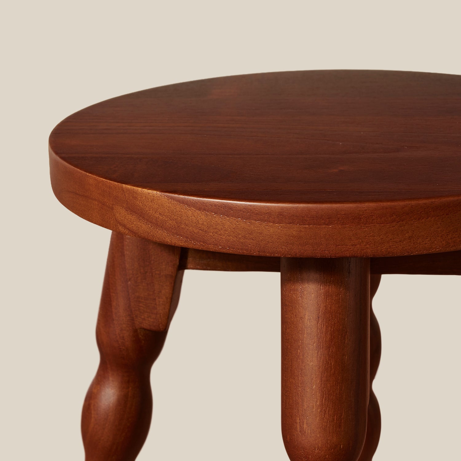 Close-up of a round wooden stool with a warm reddish-brown finish and turned wood legs, shown against a plain light beige background.- Number 4