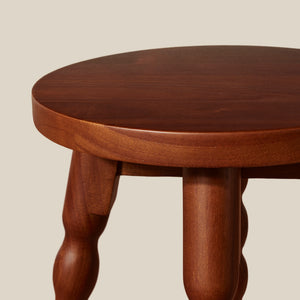 Close-up of a round wooden stool with a warm reddish-brown finish and turned wood legs, shown against a plain light beige background. - Number 4