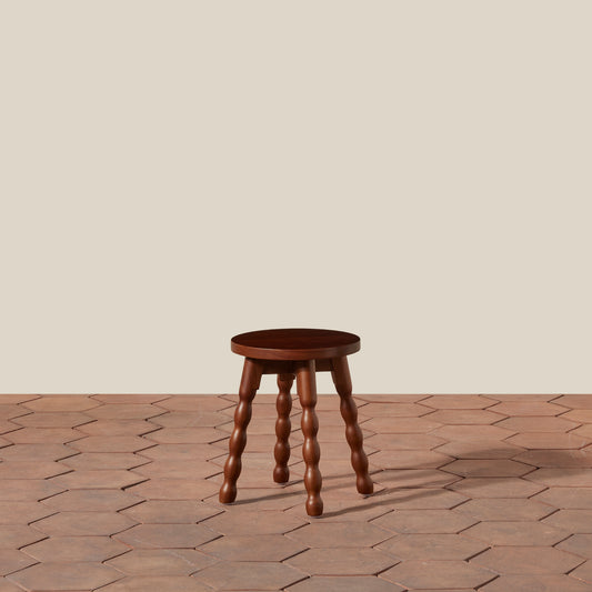 A small round wooden stool with four turned wood legs, shown on a terracotta hexagonal tile floor against a plain beige wall.