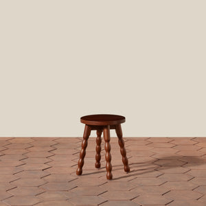 A small round wooden stool with four turned wood legs, shown on a terracotta hexagonal tile floor against a plain beige wall. - Number 1