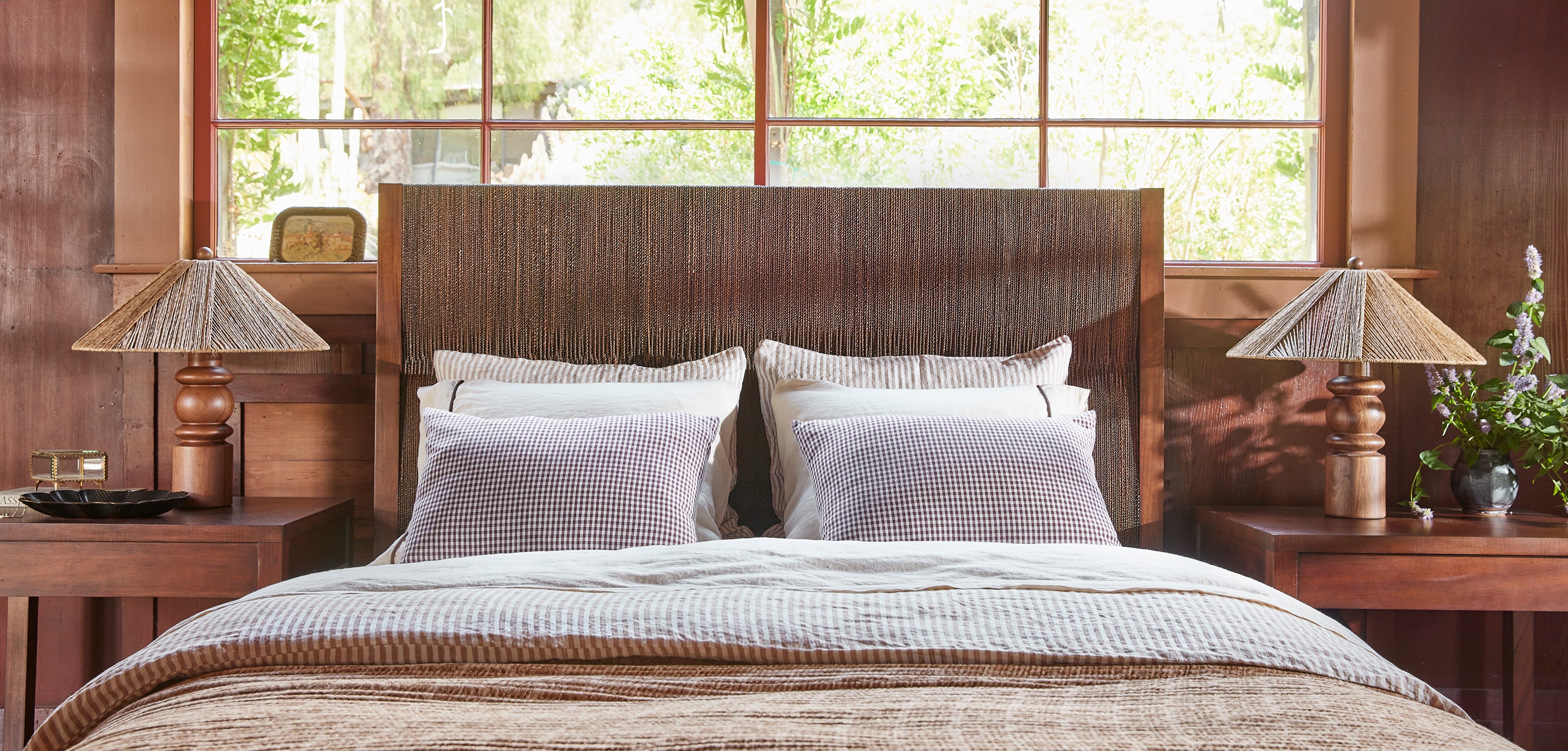 A neatly made bed with white and gray patterned pillows and a textured beige duvet, set against a tall dark wood headboard in a rustic bedroom with wooden nightstands, lamps, and a window looking out to greenery.