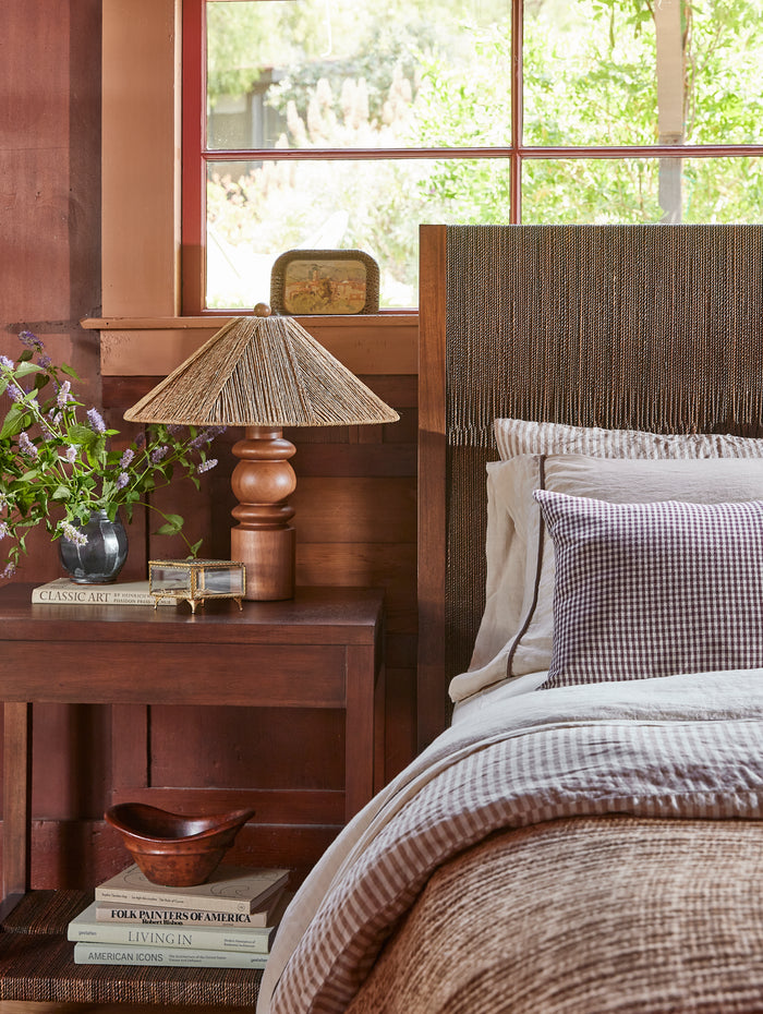 A rustic bedroom scene featuring a dark wood nightstand with a woven straw lampshade table lamp, a vase of purple flowers, and a bed with a textured brown headboard, striped bedding, and a purple-and-white gingham accent pillow.