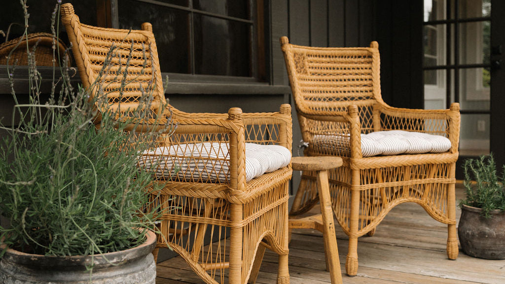 Two natural woven rattan armchairs with white cushions on a wooden porch, with a small matching side table between them and potted plants in the foreground.