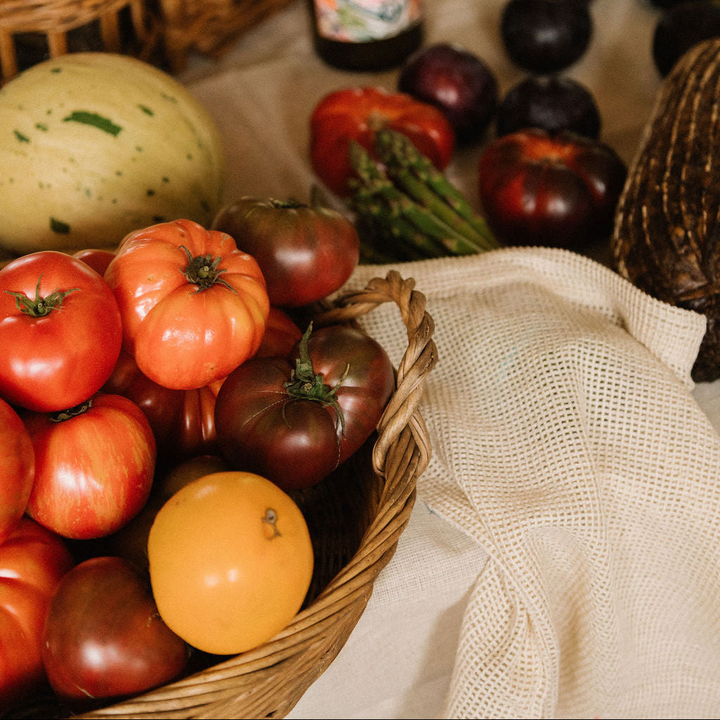 A rustic table arrangement featuring a wicker basket filled with assorted heirloom tomatoes, including red, orange, and dark purple varieties, alongside other fresh vegetables and wicker baskets in a warm, cozy setting.- Number 2