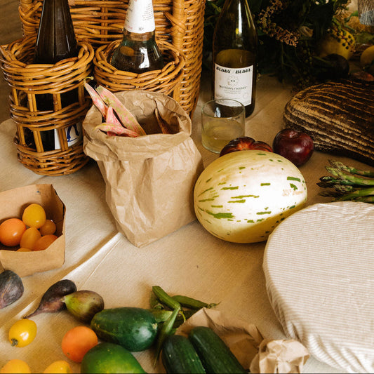 A styled picnic scene with a large woven wicker basket and matching basket holders filled with bottles, surrounded by fresh produce, cheese, and a hand holding a watermelon. A paper bag sits beside the basket on a table. - Image 2