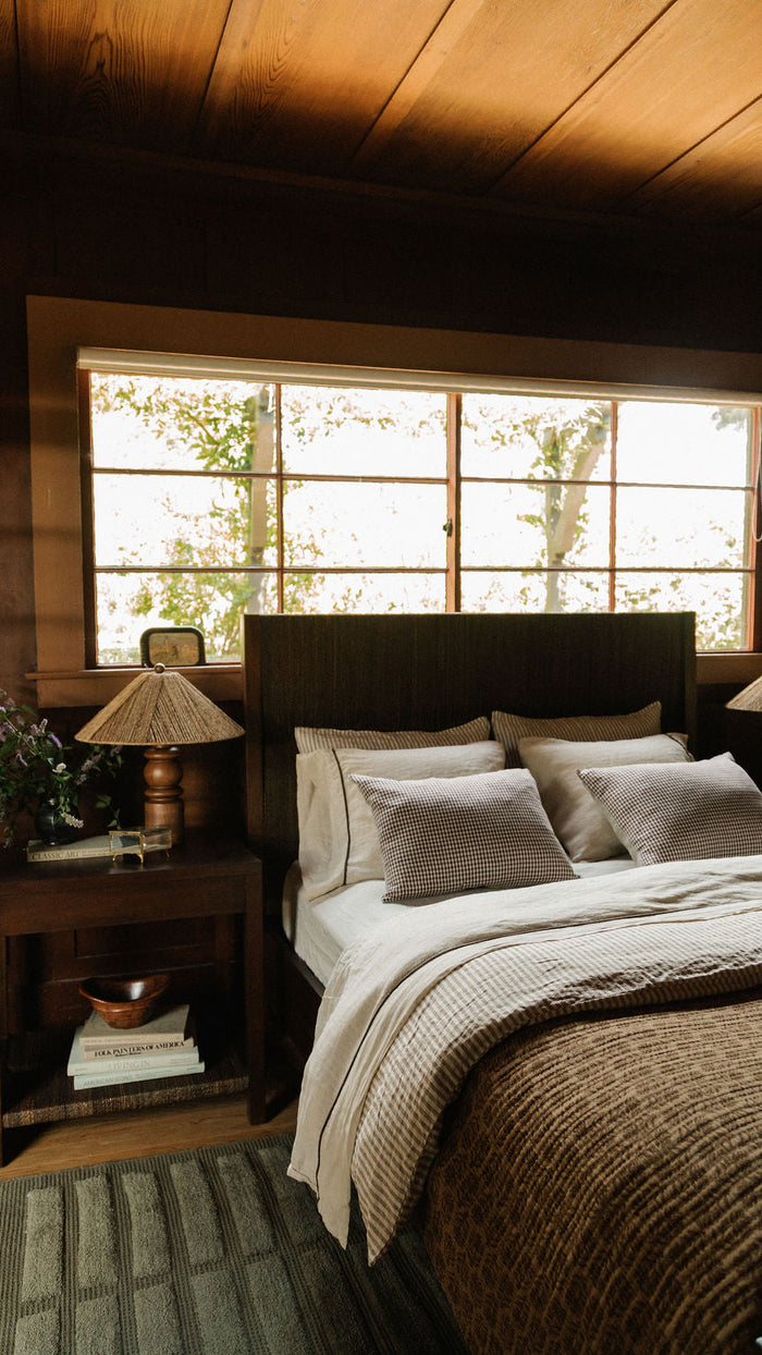 A cozy bedroom scene with a bed dressed in cream and brown textiles, set against a dark wood headboard and nightstands. A large window behind the bed shows soft outdoor light, and warm wood paneling covers the ceiling and walls.