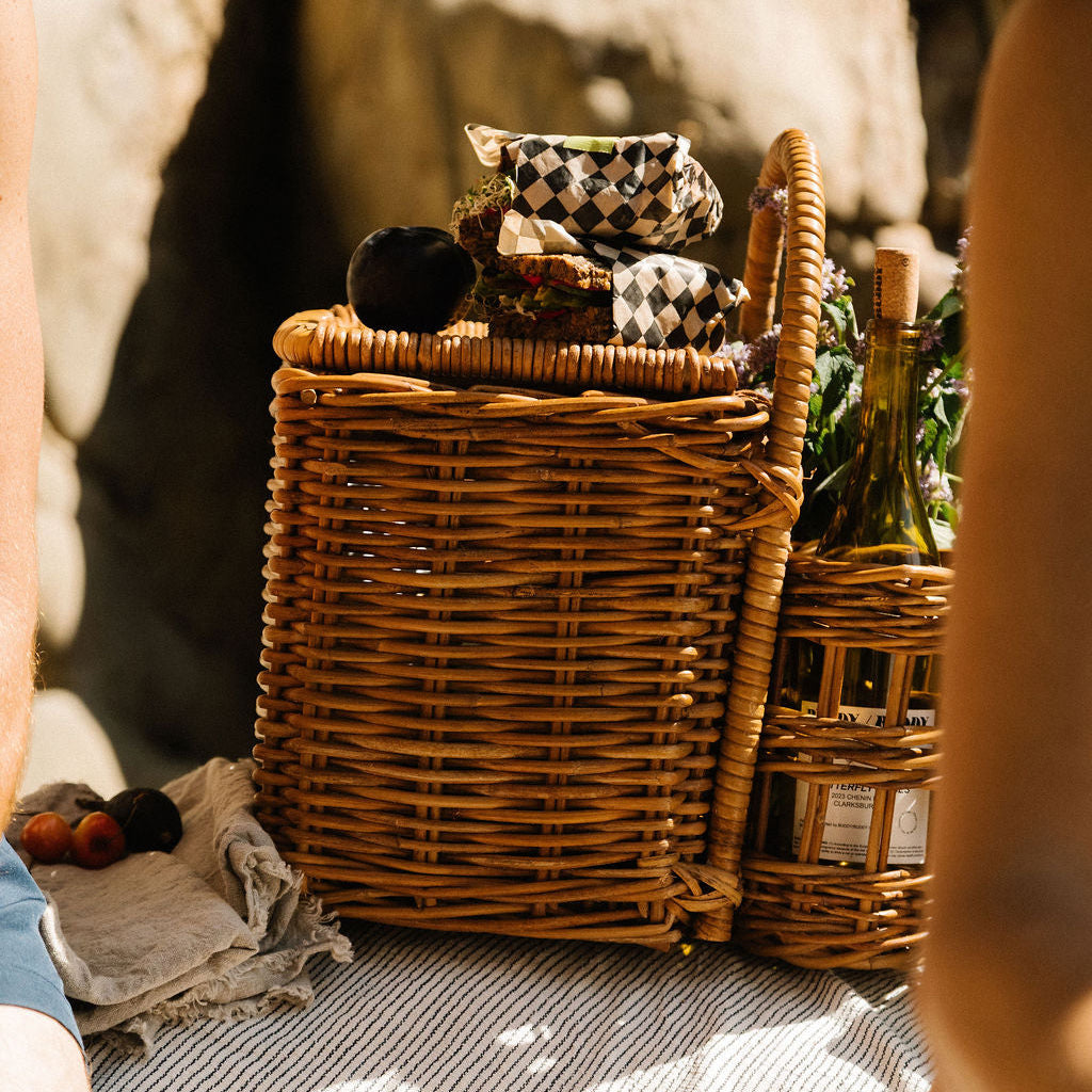 A rustic wicker picnic basket with a matching smaller side basket, filled with food and bottles, set on a striped picnic blanket outdoors with blurred human body parts in the foreground.- Number 3