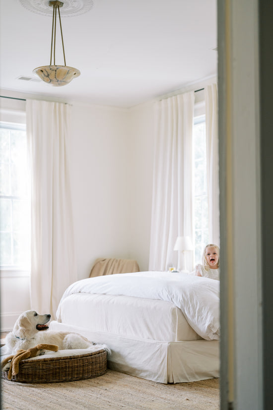 Child peeking from behind a bed in a bright bedroom with white bedding and light-colored walls.